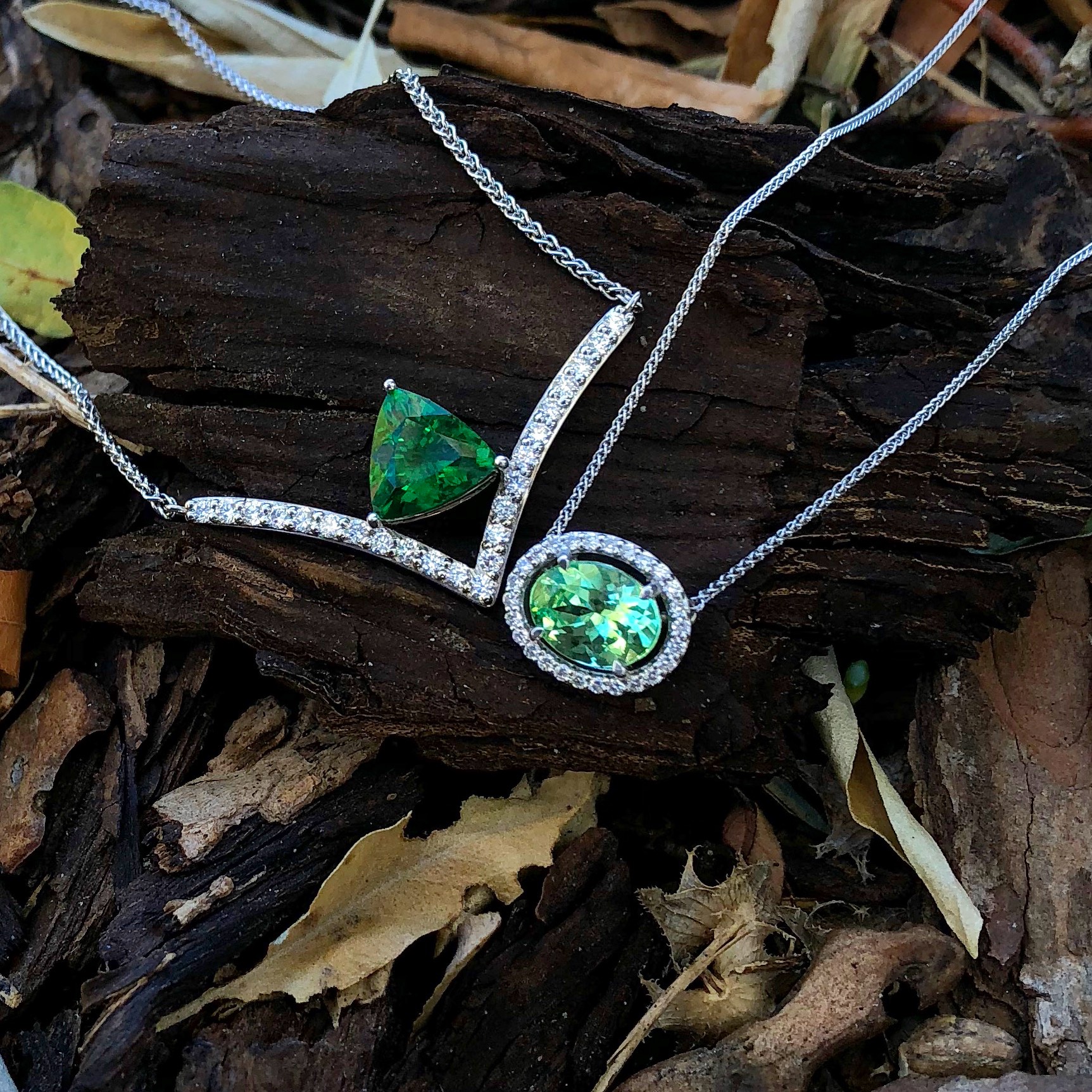 Green garnet trillion and oval pendant on a white gold chain, laid on driftwood